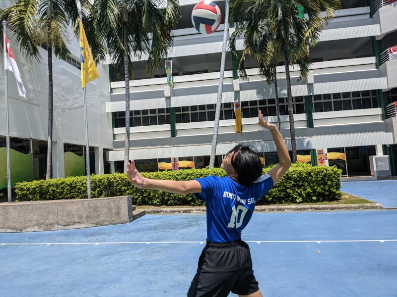 A person in a blue number 10 jersey serves a volleyball on an outdoor court near a multi-story building with palm trees.